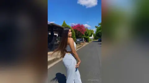 Young woman in white crop top and light jeans walking on a sunny street with blooming pink flowers.