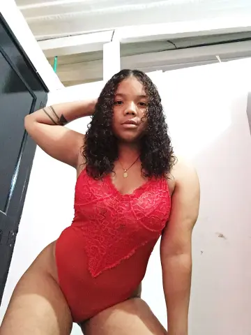 Curvy woman with curly hair wearing a red lace bodysuit in a bright indoor setting.