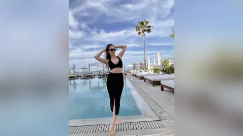 Stylish woman in a black crop top and skirt posing by a luxury resort swimming pool under a blue sky.