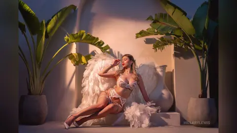 HillaryLewis poses in white lace lingerie and large white wings, sitting between two potted tropical plants in warm sunlight.