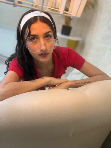Young woman with glasses and white headband wearing a red shirt, leaning on a white sofa in a bright room.