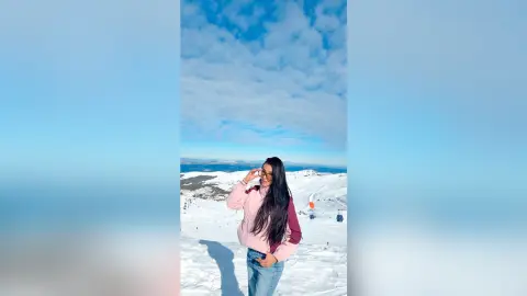 Aleja stands in a snowy mountain landscape wearing a pink puffer jacket and blue jeans.