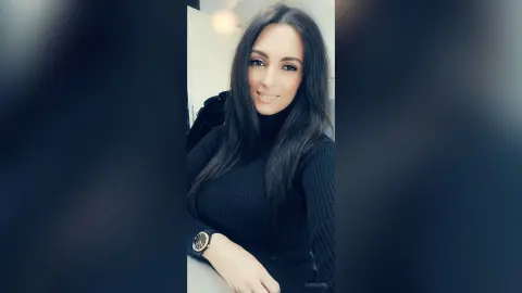 AishaApril poses in a black turtleneck, showing off her long dark hair and a gold watch on her wrist.