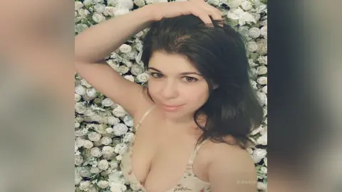 Smiling young woman in a floral camisole relaxing against a lush white rose flower wall background.