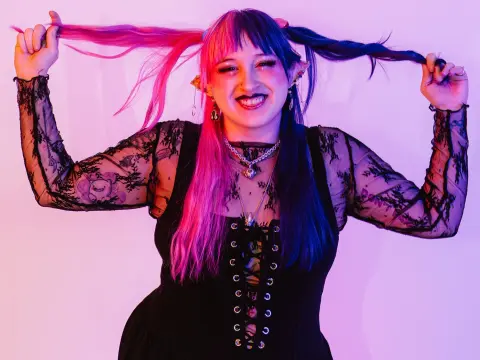 Smiling woman with pink and blue hair wearing a black lace corset in vibrant neon lighting.