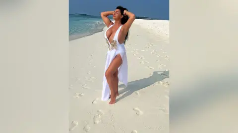 MissyJolie stands on a white sand beach wearing a white plunging-neckline dress with gold detailing.