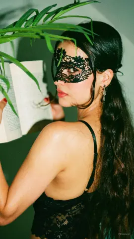 Mysterious woman in black lace mask and lingerie reading a book in a moody, tropical setting.