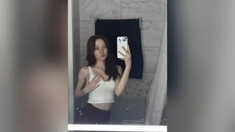 Young woman in a white ribbed tank top taking a mirror selfie in a modern marble bathroom.