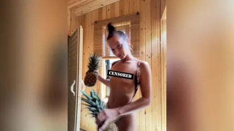 Young woman in a bikini holding a pineapple in a rustic wooden cabin setting with a relaxed summer mood.