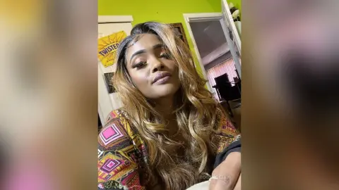 BossyBella — BossyBella poses close-up with long, wavy blonde-highlighted hair, wearing a colorful patterned top against a lime green wall.