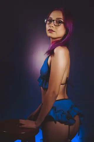 Young woman with purple hair in a blue ruffled bikini set posing in moody studio lighting.