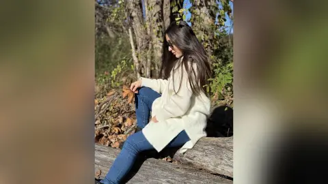 Woman in a cozy white sweater and blue jeans sitting on a log in a sunny autumn forest.
