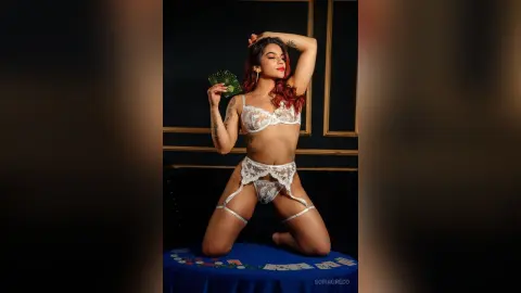 SofiaGreco poses in white lace lingerie and stockings while kneeling on a blue table and holding cash.