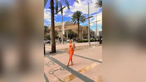 Kym stands on a paved outdoor walkway wearing a tight orange dress and red heels, surrounded by palm trees and buildings.