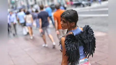 Woman in blue top and black feathered angel wings walking on a busy city street.