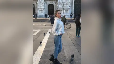 MontseFerrera stands on a stone plaza wearing a white long-sleeve top and blue jeans, looking over her shoulder.