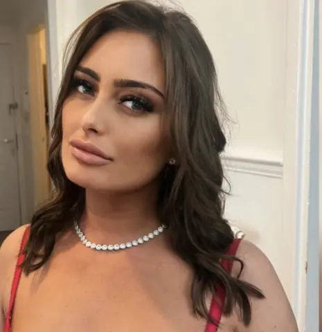 Bellamay95 poses with long wavy brown hair and a sparkling silver necklace, wearing a red top or dress.