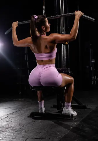 Athletic woman in pink gym wear performing lat pulldowns on a machine in a dark fitness studio.