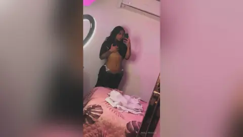 Young woman in a black crop top and dark pants taking a mirror selfie in a moody, dimly lit bedroom.
