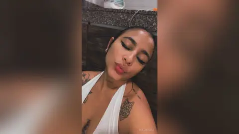 Young woman with tattoos in a white halter top posing with a relaxed expression in a modern indoor setting.