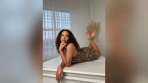 Young woman in a leopard print dress relaxing on a white bed in a bright, airy bedroom.