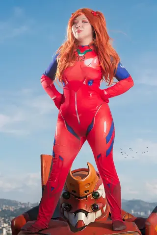 Redheaded woman in a red Evangelion Unit 02 bodysuit standing on a robot prop under a blue sky.