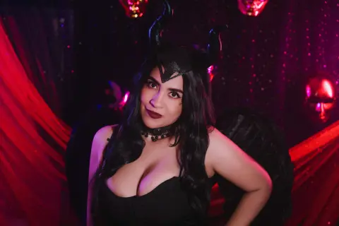 Woman in dark devil costume with black horns and wings posing in a moody red lit setting.