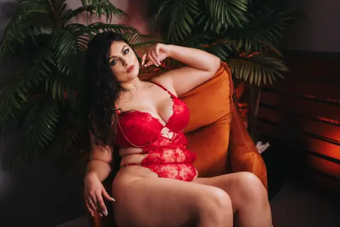 Curvy woman in red lace lingerie relaxing on an orange armchair in a moody, tropical indoor setting.