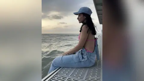 CrissRusso sits on a metal deck overlooking the ocean, wearing a light blue cap, a pink bikini top, and light blue jeans.