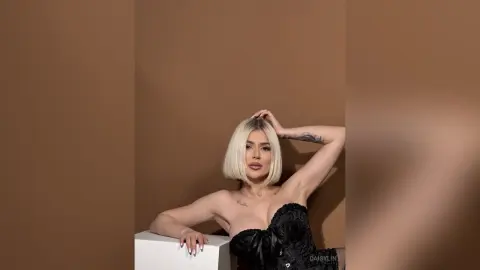 Glamorous blonde woman in a black lace corset posing against a brown studio background.