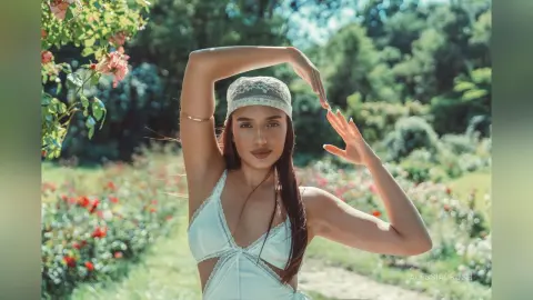 AlessiaCrush poses in a white lace outfit and headscarf while standing in a sunny garden filled with flowers.