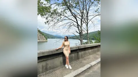 Stylish woman in a beige midi dress leaning on a stone wall by a scenic mountain lake under a cloudy sky.