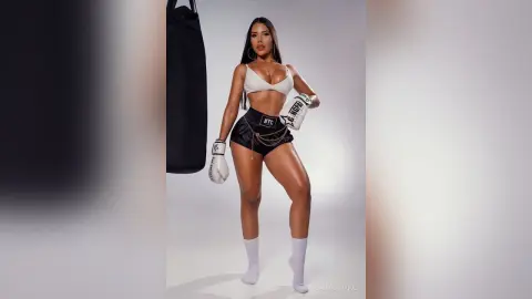 CassandraDuque poses in white boxing gloves, a white bra, and black shorts against a plain background.
