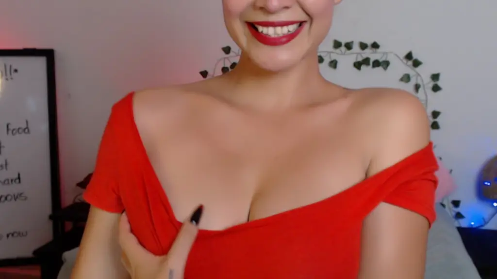 SofiaPardo smiles while wearing a pink floral strapless top, showing her red hair and shoulders while sitting on a bed with string lights behind her.