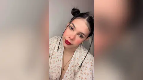 Young woman with dark hair in a top knot wearing a floral print blouse and red lipstick in a bright setting.