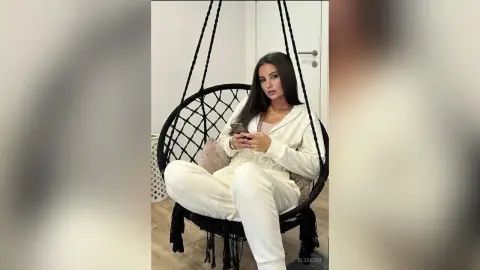Young woman in cozy white loungewear relaxing in a black hanging egg chair in a bright modern room.