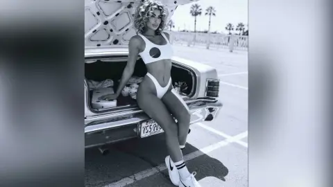 SinnaRay poses in a white bikini by the open trunk of a vintage car on a sunny day.