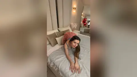 Young woman in a pink top relaxing on a white bed in a bright, modern bedroom setting.