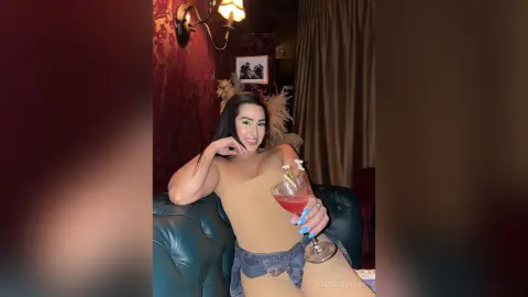 Glamorous woman in a beige bodysuit holding a cocktail in a moody, dimly lit lounge setting.