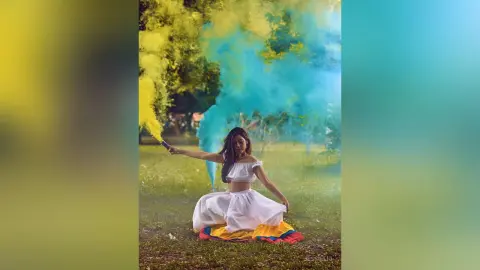 JessieLiu poses in a white off-the-shoulder top and skirt while holding a yellow smoke flare against a blue smoke background.