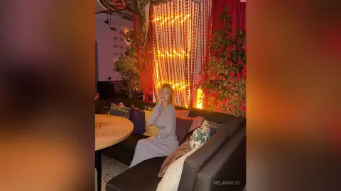 Woman in a grey dress relaxing on a sofa in a cozy, warm-lit room with fairy lights and indoor plants.