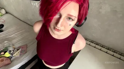 Young woman with red hair and facial piercings wearing a maroon tank top in a bright bedroom setting.