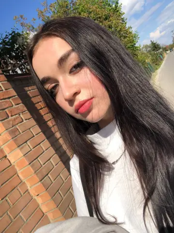 Young woman with long dark hair wearing a white turtleneck outdoors in bright sunlight.