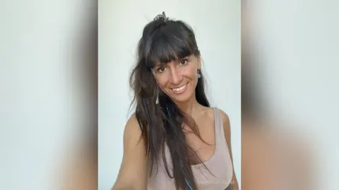 Smiling brunette woman with long hair in a casual beige tank top posing against a white background.