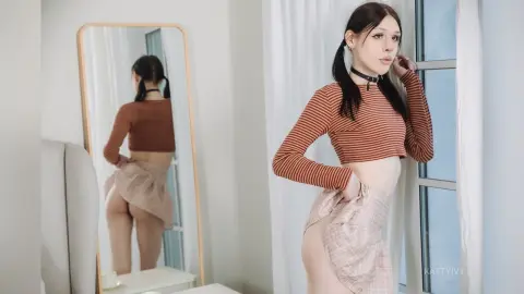 Young woman in a brown striped crop top and pink mini skirt posing by a window in a bright room.