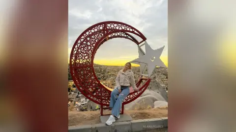 AlanaPearce sits inside a large red crescent moon and star sculpture, wearing a light tan jacket and blue jeans against a sunset sky.