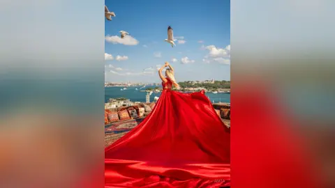 Anita poses with her arms raised in a long red gown against a blue sky and ocean backdrop.