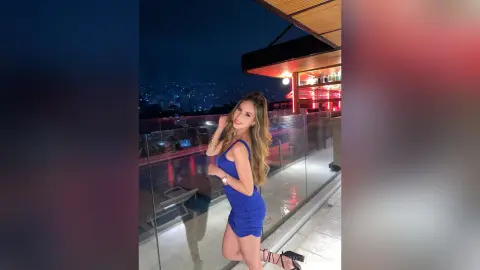 Smiling woman in a blue cocktail dress posing on a rooftop balcony overlooking a city skyline at night.