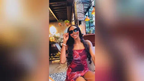 Smiling woman in a red floral dress and sunglasses enjoying a sunny outdoor cafe setting.
