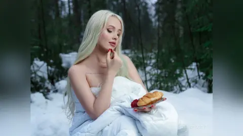 EllieFrosty with long platinum blonde hair holds a plate of pastries and a red berry while wrapped in a white duvet in a snowy forest.
